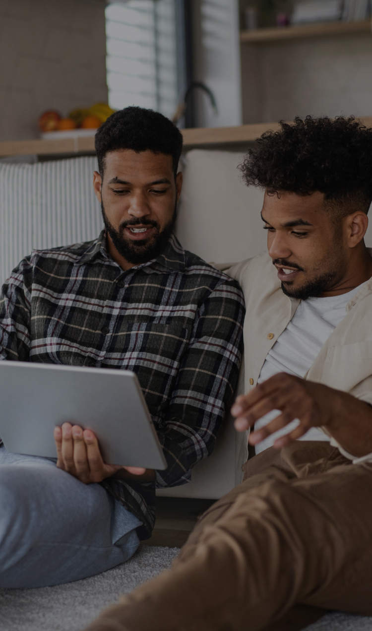 A father and son sit together on the floor of the family room while attending a virtual family therapy session on a tablet.